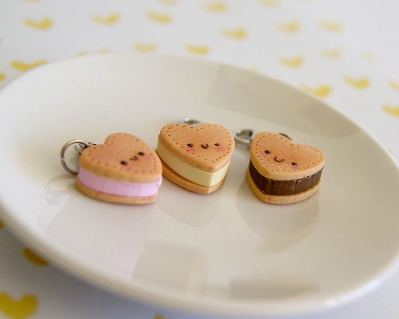 May include: Three heart-shaped keychains resembling ice cream sandwiches. Each keychain has a light brown cookie exterior with a different colored filling: pink, yellow, and brown. The keychains have smiling faces and are on a white plate.