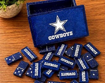 May include: A blue domino set with white dots and the word "COWBOYS" printed on each piece. The set includes a matching box with the Cowboys star logo. The dominoes are scattered on a wooden surface.