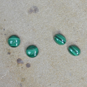 May include: Four green malachite cabochons, oval and round shaped, on a light brown surface.