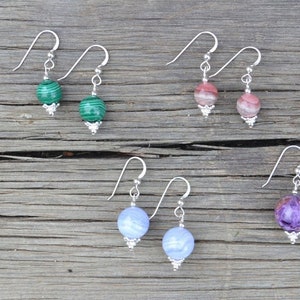 May include: A collection of gemstone earrings, each with a silver hook and a decorative silver bead. The gemstones include malachite, rhodochrosite, blue lace agate, and charoite. The earrings are displayed on a wooden surface.
