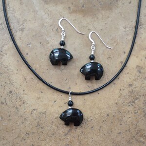 K&ouml;nnte beinhalten: Eine Halskette mit einem Anh&auml;nger in Form eines B&auml;ren aus schwarzem Onyx und dazu passende Ohrringe an einem schwarzen Lederband.