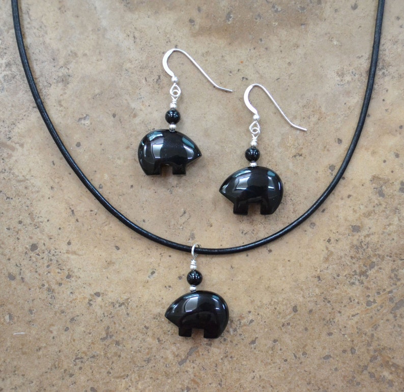 K&ouml;nnte beinhalten: Eine Halskette mit einem Anh&auml;nger in Form eines B&auml;ren aus schwarzem Onyx und dazu passende Ohrringe an einem schwarzen Lederband.