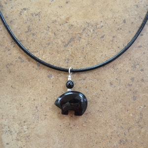 K&ouml;nnte beinhalten: Eine Halskette mit einem schwarzen Onyx-B&auml;renanh&auml;nger an einem schwarzen Lederband. Der Anh&auml;nger ist in einer einfachen, stilisierten B&auml;renform gehalten.