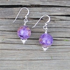 May include: A pair of dangle earrings featuring round, purple and brown marbled beads. The beads are suspended from silver-colored hooks and have decorative silver-colored accents at the base. The earrings are displayed on a weathered wooden surface.