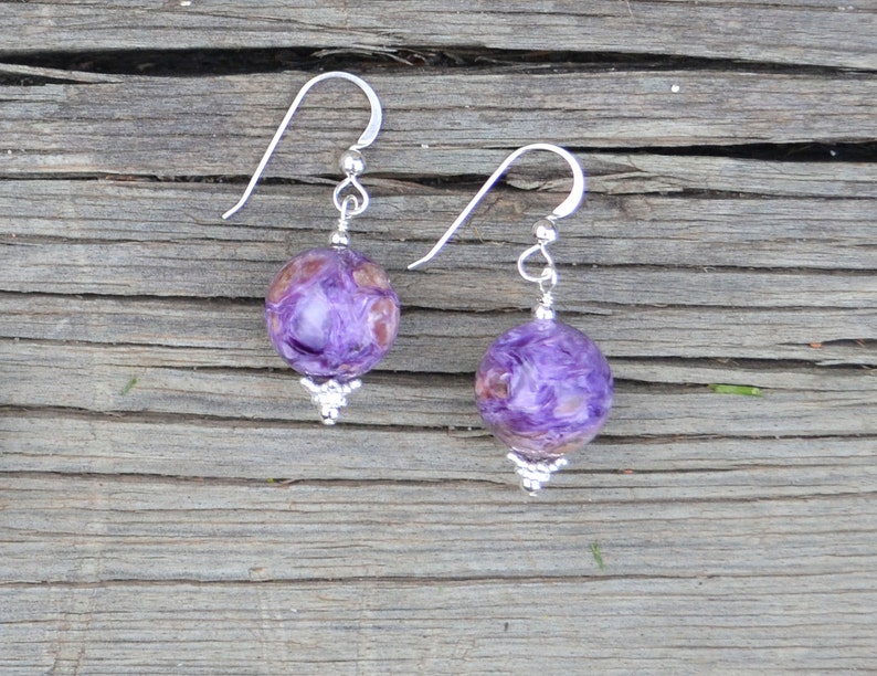 May include: A pair of dangle earrings featuring round, purple and brown marbled beads. The beads are suspended from silver-colored hooks and have decorative silver-colored accents at the base. The earrings are displayed on a weathered wooden surface.