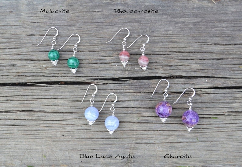 May include: A collection of gemstone earrings, each with a silver hook and a decorative silver bead. The gemstones include malachite, rhodochrosite, blue lace agate, and charoite. The earrings are displayed on a wooden surface.