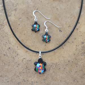 May include: A black leather necklace with a silver turtle pendant and matching earrings. The turtle is decorated with colorful geometric patterns in shades of blue, green, orange, and red.