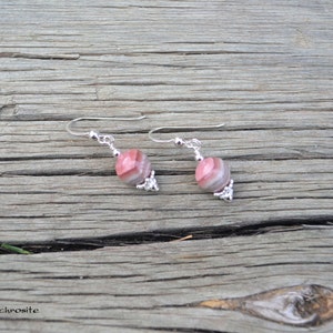 May include: A pair of earrings featuring pink and gray striped beads with silver-colored accents. The earrings are displayed on a weathered wooden surface. The word "Rhodochrosite" is visible in the lower left corner.