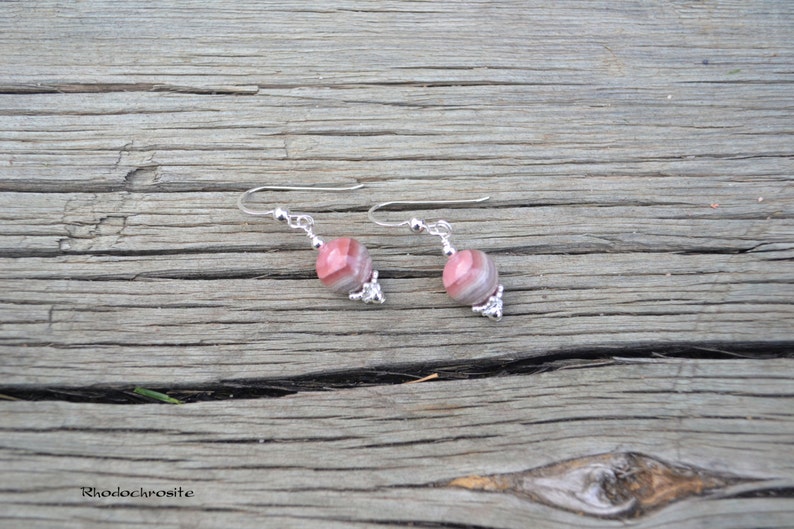 May include: A pair of earrings featuring pink and gray striped beads with silver-colored accents. The earrings are displayed on a weathered wooden surface. The word "Rhodochrosite" is visible in the lower left corner.