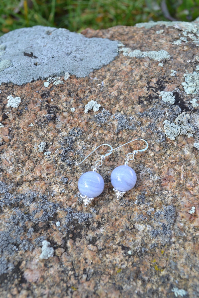 May include: A pair of light blue, round gemstone earrings with silver-colored hooks and decorative accents. The earrings are displayed on a textured, gray and brown rock surface, with green grass visible in the background. The earrings are a type of jewelry.