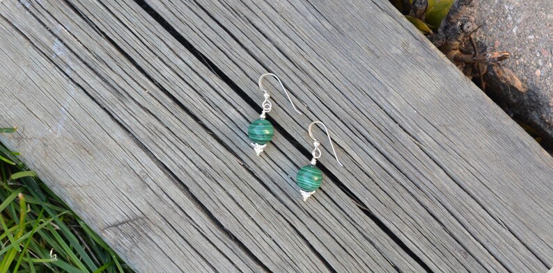May include: A pair of silver earrings with green malachite beads. The earrings have a simple hook design and a decorative silver cap at the base of each bead. The earrings are displayed on a weathered wooden surface.