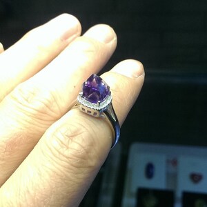 Puede incluir: Un anillo de tono plateado que presenta una piedra preciosa de amatista de color p&uacute;rpura intenso y facetada en el centro. La piedra est&aacute; rodeada por un delicado halo de peque&ntilde;as piedras claras y brillantes engastadas en la banda de metal pulido.