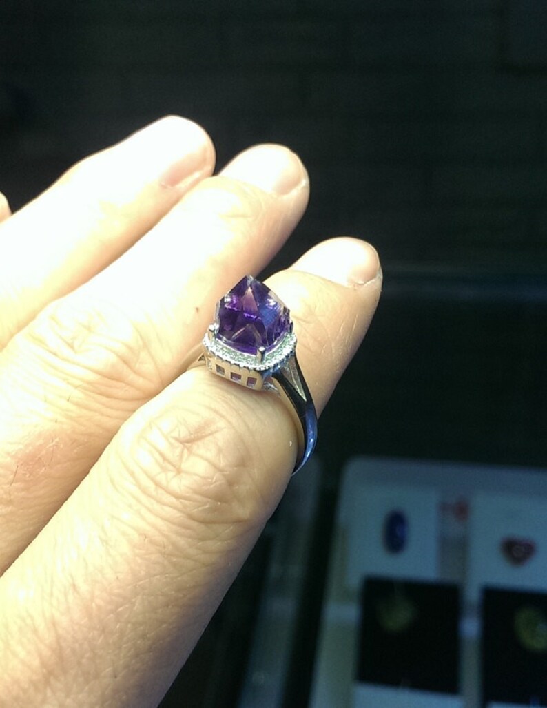 Puede incluir: Un anillo de tono plateado que presenta una piedra preciosa de amatista de color p&uacute;rpura intenso y facetada en el centro. La piedra est&aacute; rodeada por un delicado halo de peque&ntilde;as piedras claras y brillantes engastadas en la banda de metal pulido.