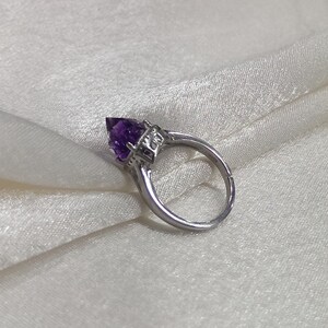 Puede incluir: Un anillo de tono plateado con una piedra preciosa amatista morada facetada en forma de pera, montada en un engaste de garras. La banda de metal tiene una abertura visible en la parte posterior, lo que indica un dise&ntilde;o ajustable y decorativo.