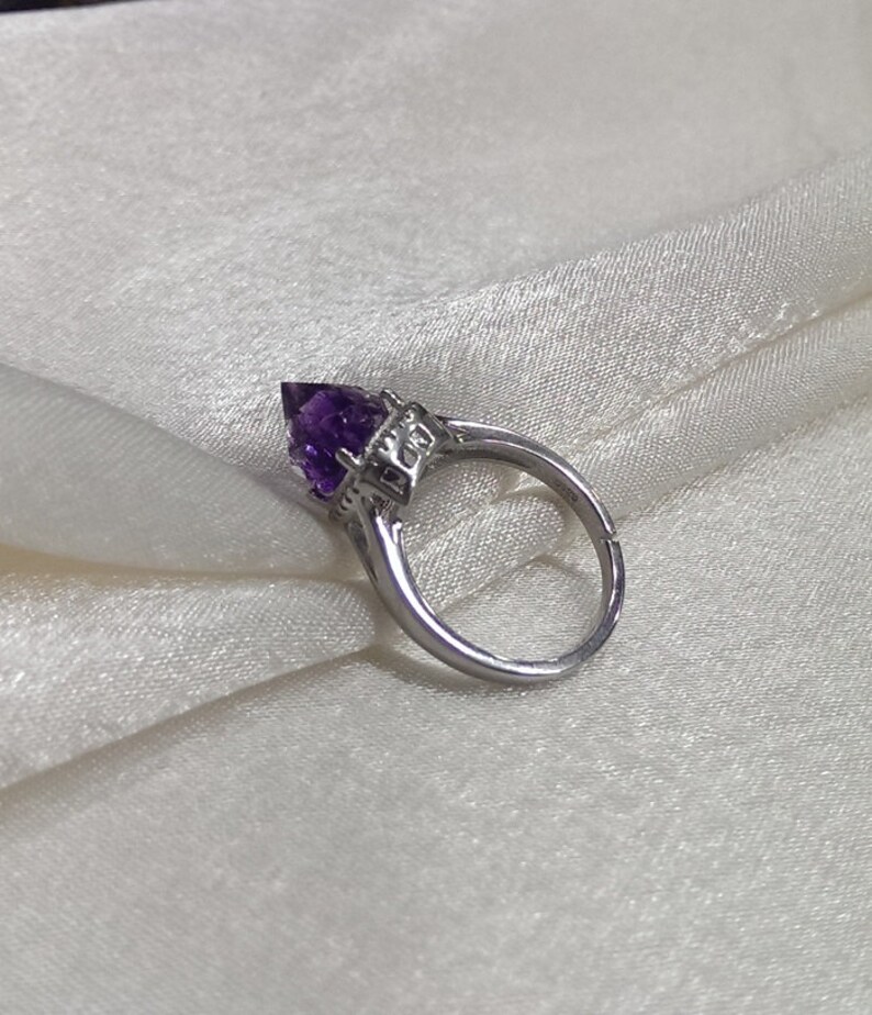 Puede incluir: Un anillo de tono plateado con una piedra preciosa amatista morada facetada en forma de pera, montada en un engaste de garras. La banda de metal tiene una abertura visible en la parte posterior, lo que indica un dise&ntilde;o ajustable y decorativo.