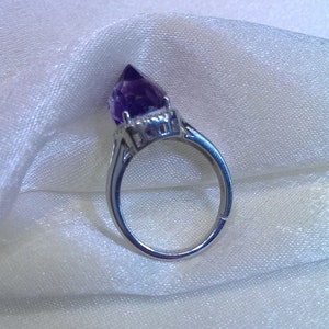 Puede incluir: Un anillo de tono plateado con un cristal de amatista de color p&uacute;rpura intenso y forma puntiaguda, montado en una base de metal con garras. La banda es lisa y met&aacute;lica, mostrando una piedra preciosa facetada que se eleva en una punta afilada.