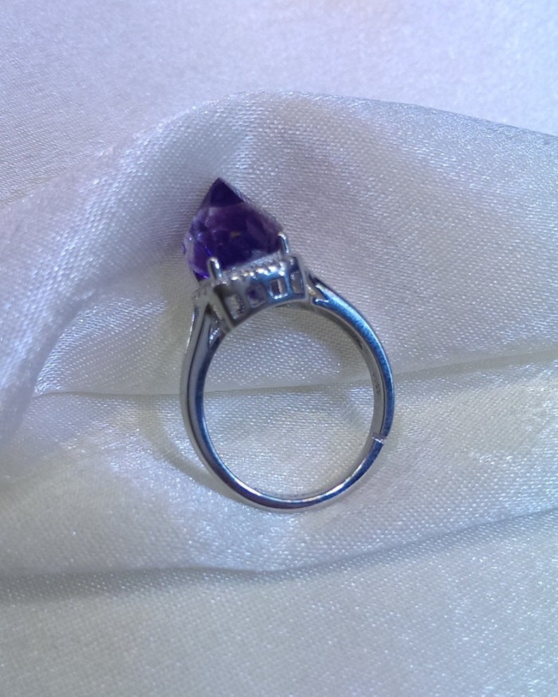 Puede incluir: Un anillo de tono plateado con un cristal de amatista de color p&uacute;rpura intenso y forma puntiaguda, montado en una base de metal con garras. La banda es lisa y met&aacute;lica, mostrando una piedra preciosa facetada que se eleva en una punta afilada.