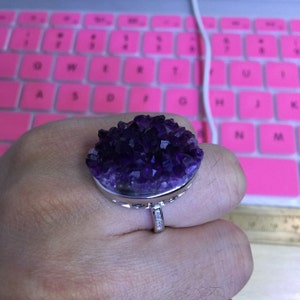 Puede incluir: Un anillo de tono plateado con una gran piedra central circular de drusa de amatista de color púrpura intenso. La banda está decorada con pequeñas piedras claras y brillantes en el lateral, creando un contraste con la superficie de cristal oscura y texturizada.