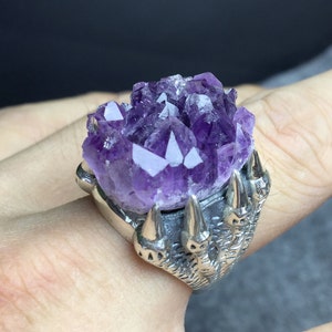 Puede incluir: Un anillo de tono plateado con un gran racimo de cristales de amatista púrpura en bruto, engastado en una banda de metal diseñada para parecer una garra. El engaste tiene cuatro puntas afiladas y distintas que sujetan la base de la gema.