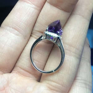 Puede incluir: Un anillo de tono plateado con una gema de amatista cruda de color p&uacute;rpura intenso, engastada en una cesta de metal con garras. La banda es lisa y met&aacute;lica, mostrando las facetas naturales, sin pulir y angulares de la piedra de cristal.
