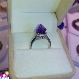 Puede incluir: Un anillo de tono plateado con una punta de cristal de amatista en bruto de color violeta intenso como pieza central. El engaste incluye un halo de peque&ntilde;as piedras claras que rodean la base del cristal, montado sobre una banda de metal pulido.