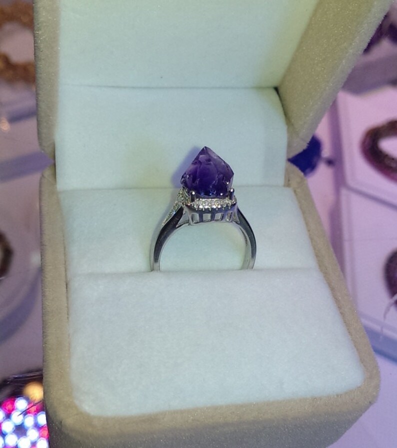 Puede incluir: Un anillo de tono plateado con una punta de cristal de amatista en bruto de color violeta intenso como pieza central. El engaste incluye un halo de peque&ntilde;as piedras claras que rodean la base del cristal, montado sobre una banda de metal pulido.