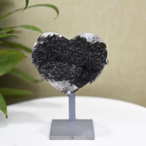 May include: A heart-shaped black crystal geode on a gray metal stand. The geode has a rough, textured surface with a dark, granular interior and white edges. The stand has a square base and a vertical support.
