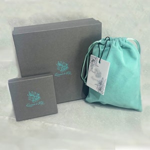May include: Two gray boxes with the text "Queens & Lily" in teal on the front. The larger box is closed and the smaller box is open. A teal velvet bag with a white tag is in front of the boxes.