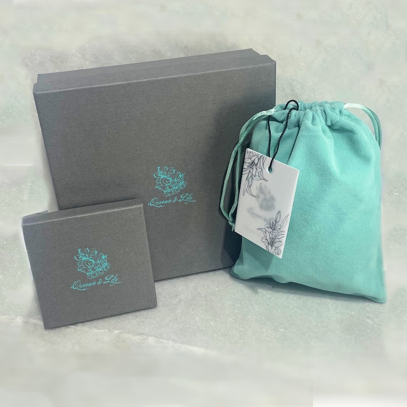 May include: Two gray boxes with the text "Queens & Lily" in teal on the front. The larger box is closed and the smaller box is open. A teal velvet bag with a white tag is in front of the boxes.