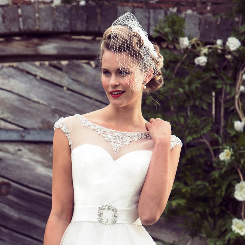 May include: A white wedding dress with a sweetheart neckline and a sheer bodice with beaded embellishments. The dress has a fitted bodice and a full skirt. The bride is wearing a white birdcage veil and a jeweled belt.