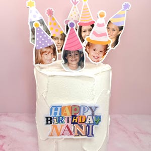 Pode incluir: Um bolo com cobertura branca decorado com um topo de bolo personalizado com várias fotografias recortadas de rostos usando chapéus de festa coloridos. Uma placa retangular na frente do bolo exibe o texto HAPPY BIRTHDAY NANI em letras coloridas.