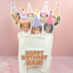 Pode incluir: Um bolo com cobertura branca apresentando um topo de bolo personalizado com o texto HAPPY BIRTHDAY NANI em letras laranja. O topo inclui vários recortes de rostos usando chapéus de festa ilustrados em várias cores.