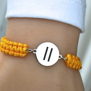 May include: A yellow braided bracelet with a silver round charm featuring two parallel lines.