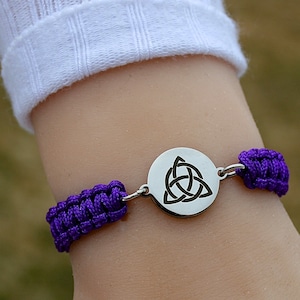 May include: A purple macrame bracelet with a silver circular charm featuring a Celtic knot design. The bracelet is connected with silver metal links. The bracelet is worn on a wrist.