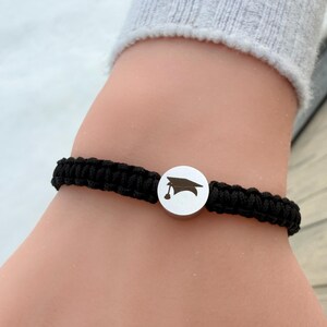 May include: Black braided bracelet with a silver charm depicting a graduation cap. The bracelet is worn on a wrist, highlighting the contrast between the dark cord and the bright charm.