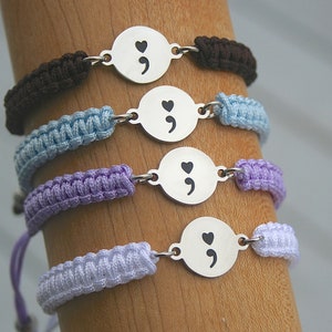 May include: Four macrame bracelets in brown, light blue, purple, and lavender. Each bracelet features a round, silver-toned charm with a semicolon and heart symbol. The bracelets are displayed on a wooden dowel.