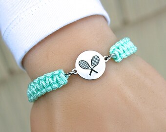 May include: A tennis-themed bracelet featuring a round silver charm with crossed tennis rackets. The charm is connected to light turquoise woven bands. The bracelet is designed to be worn on the wrist.