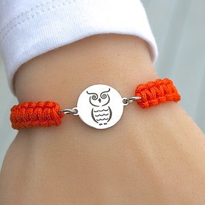 May include: A silver bracelet featuring an owl design. The bracelet has a round, engraved pendant with an owl illustration. The bracelet is connected by bright orange woven cords.
