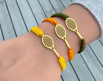 May include: Three tennis racket charm bracelets. The bracelets feature gold-colored tennis racket charms attached to braided cords in yellow, orange, and green. The bracelets are displayed on a person's wrist.