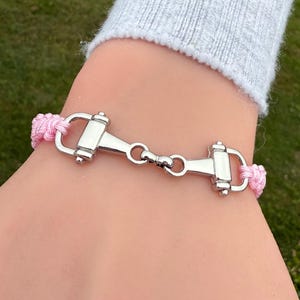 May include: A pink braided bracelet with a silver-colored equestrian bit charm. The bracelet is worn on a wrist, with a gray long-sleeved shirt in the background. The bracelet is a stylish accessory.