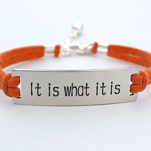 Peut inclure: Bracelet en daim orange avec une plaque rectangulaire argentée gravée de la phrase "It is what it is". Le bracelet est doté d'un fermoir argenté et est réglable.