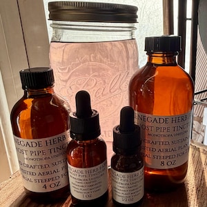 May include: An assortment of amber glass bottles with black droppers and labels, containing herbal tinctures. A large Ball jar with a metal lid and pink liquid is also present. The labels read "Enegade Herb Farm" and list the ingredients and size.