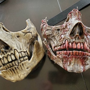Puede incluir: Dos máscaras de calavera de media cara. Una es de color hueso desgastado, la otra es de tono de carne en descomposición con dientes expuestos y detalles similares a la sangre. Ambas máscaras tienen correas negras para asegurar.