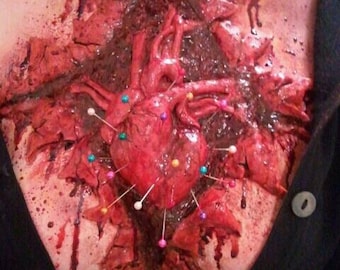 May include: A gruesome Halloween costume featuring a realistic, open-chest wound with a visible heart. The heart is adorned with colorful pins, and the wound is detailed with blood and gore. The costume is worn over a black shirt.