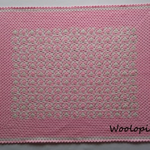 Puede incluir: Una manta de beb&eacute; de crochet rosa y blanca con un patr&oacute;n geom&eacute;trico. La manta tiene un borde blanco y el texto "Woolop&iacute;a" es visible en la esquina inferior derecha.