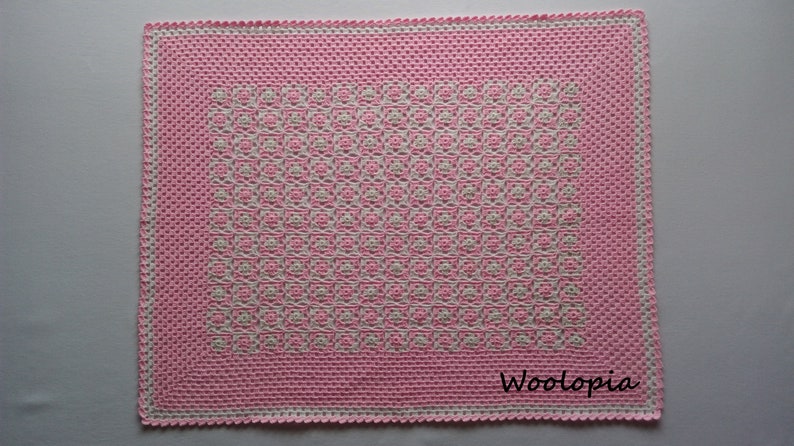 Puede incluir: Una manta de beb&eacute; de crochet rosa y blanca con un patr&oacute;n geom&eacute;trico. La manta tiene un borde blanco y el texto "Woolop&iacute;a" es visible en la esquina inferior derecha.
