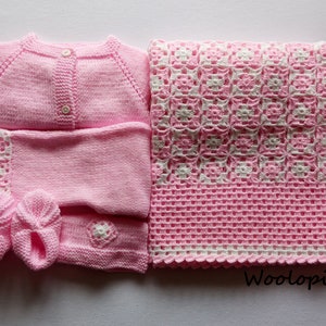 Puede incluir: Un c&aacute;rdigan de punto rosa para beb&eacute;, pantalones a juego, botines y una manta de ganchillo rosa y blanca. La manta tiene un borde blanco y un patr&oacute;n repetitivo de flores blancas sobre un fondo rosa. El texto "Woolop&iacute;a" es visible en la esquina inferior derecha de la imagen.
