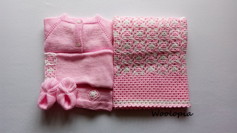 Puede incluir: Un c&aacute;rdigan de punto rosa para beb&eacute;, pantalones a juego, botines y una manta de ganchillo rosa y blanca. La manta tiene un borde blanco y un patr&oacute;n repetitivo de flores blancas sobre un fondo rosa. El texto "Woolop&iacute;a" es visible en la esquina inferior derecha de la imagen.