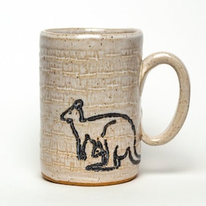 Könnte beinhalten: Eine Keramiktasse mit beigefarbener Glasur und einer schwarzen Umrandung eines Kängurus mit einem Joey in seiner Tasche. Die Tasse hat eine strukturierte Oberfläche und einen abgerundeten Griff.