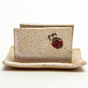 May include: A ceramic napkin holder with a ladybug design. The holder is white with brown speckles and a leaf pattern. It has a matching tray.